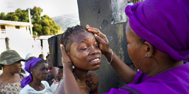 PORT-AU-PRINCE, HAITI - NOVEMBER 1, 2010: A Vodou priestess, or mambo, baths a young woman to facilitate her passage into the spirit world in front of a cross dedicated to Baron Samedi, the head of the Gede family of spirits, the spirits of the dead, in the National Cemetery in Port-au-Prince, Haiti, on All Saints Day, November 1, 2010. The cross is a symbol of the crossroad between the living and the dead. All Saints Day and the Day of the Dead are two days devoted to the Gede, who are feted for most of the month of November. One of the most important Vodou holidays in Haiti, Vodouists go to cemeteries to pray with food, coffee and peppered alcohol, to light candles and to put fresh flowers on graves, and then dance all night at 'peristyles' or Vodou temples. (Photo by Dario Mitidieri/Getty Images)