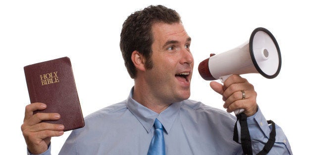 Young caucasian male yelling into megaphone while holding Holy Bible in other hand. He is wearing a blue shirt and blue tie.