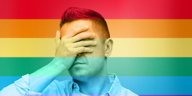 homosexual, homophobia, intolerance and people concept - unhappy gay man covering his eyes by hand over rainbow flag backgrou