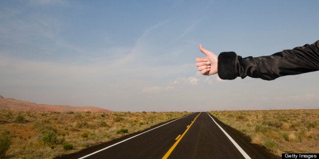 An elegantly dressed womans arm and hand are held out hitch hiking on a deserted road.