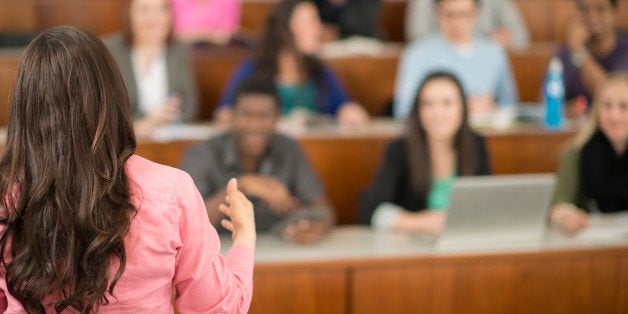 A multi-ethnic group of college age students are sitting in a row in a lecture hall and are listening to their professor.