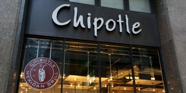 A Chipotle eatery in the Financial District of Manhattan NYC viewed from the outside looking in shows the interior design as well as people eating & talking, & also reflects the urban building from across the street in the upper left corner of the window