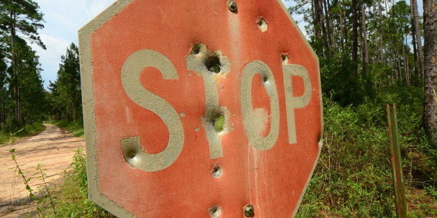 Angled photo of an old 'STOP' sign in a forest, with faded paint and various sized bullet holes. A dirt road at the left of t