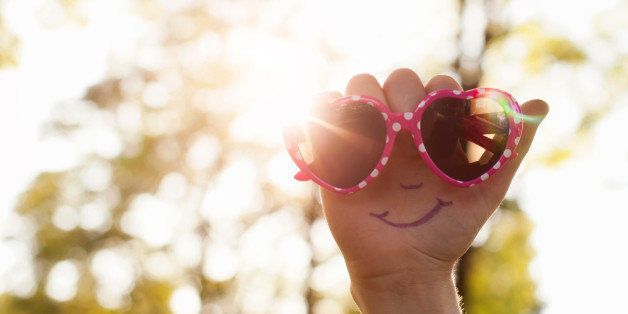 Hand with a smiling face drawn on palm, holding a pair of heart shaped sunglasses up to the sun.