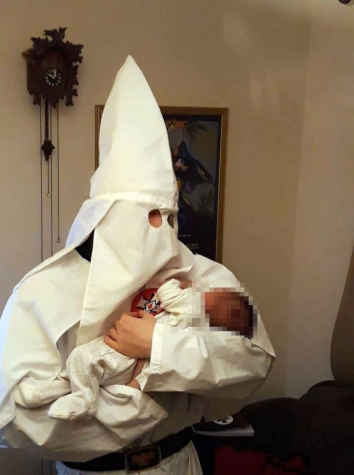 Adam Thomas wearing the hooded white robe of a Ku Klux Klansman, holding his newborn baby at home