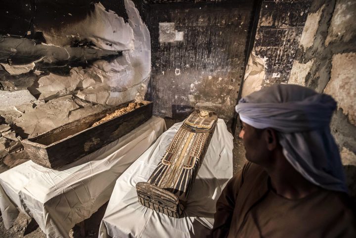 An Egyptian worker standing next to an opened intact sarcophagus containing a well-preserved mummy of a woman named "Thuya" wrapped in linen.