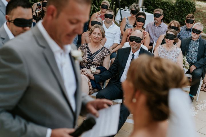 Wedding guests wear black blindfolds during the ceremony. One woman in the center of the shot is not wearing a blindfold: Agnew's mom, who also has cone-rod dystrophy. In the foreground and out of focus, you can see the groom in a gray suit and the bride in a veil with her hair pulled back in a chignon.