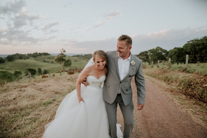 The bride and groom stand together laughing on a dirt road with green hills and a pale blue, partly cloudy sky behind them. The groom has his arm wrapped around the bride's waist as she holds her skirt in one hand.