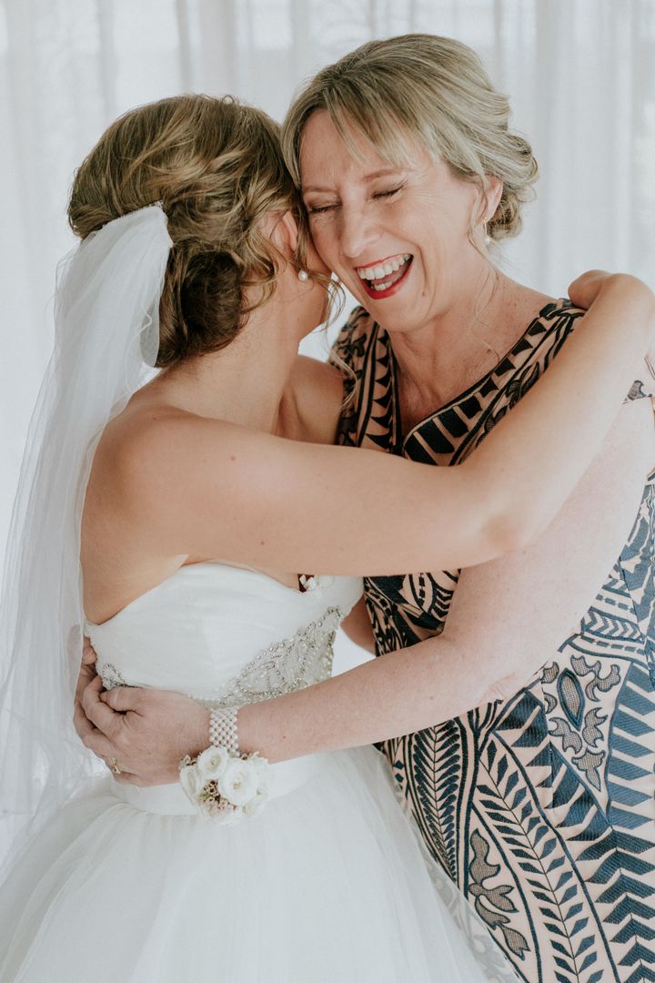 The bride and her mother share a joyful embrace. On the left is Agnew in a white ball gown and veil, with her head turned away from the camera. On the right is Agnew's mother with a big smile on her face, wearing a tan-and-navy patterned dress.