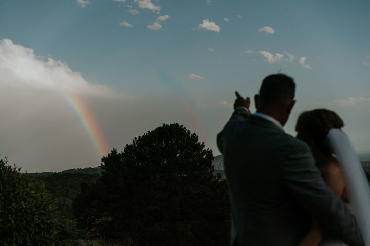 A silhouette shot of the bride and groom outdoors. The groom has his right arm wrapped around the bride as he points to a rainbow in the distance. 