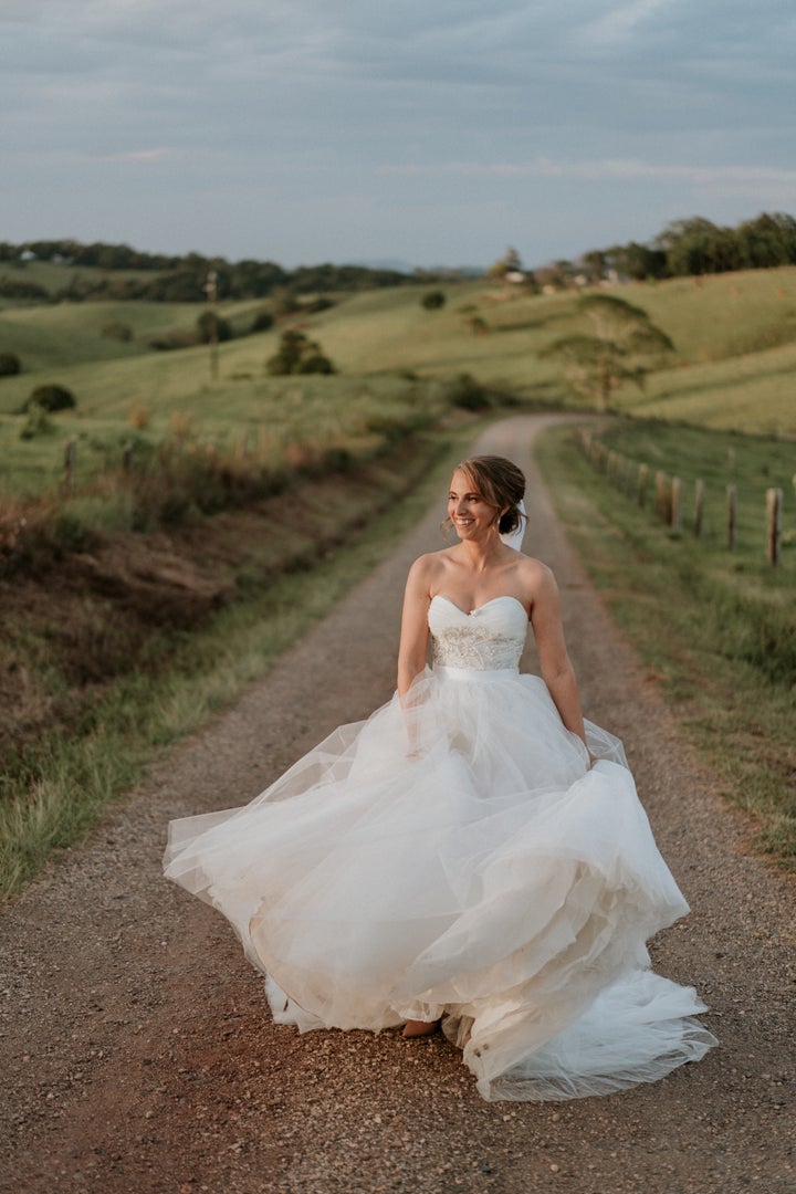 The bride, standing on a dirt road between green rolling hills, smiles as she plays with the skirt of her ball gown. 