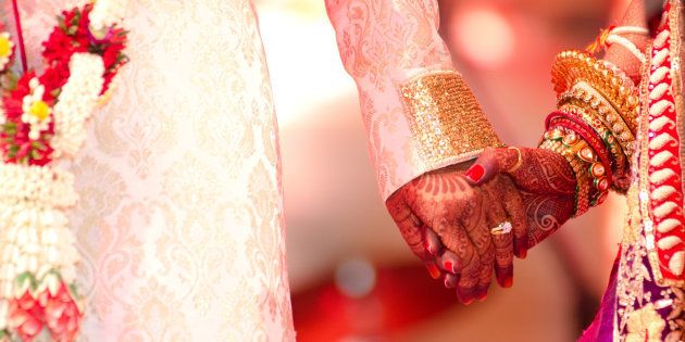 Groom holding henna decorated hand of bride laden with beautiful gold jewelry.