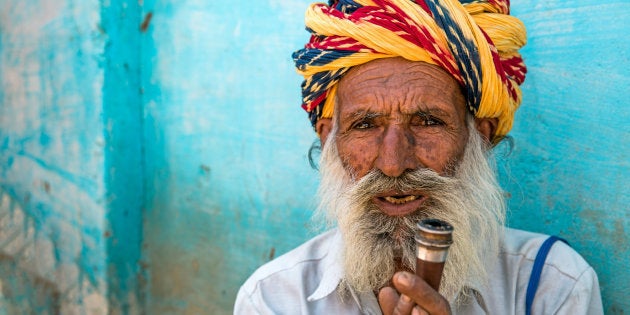 Senior indian man with vibrant colorful turban holding his tobacco pipe in front of blue wall in the streets of Rajasthan.