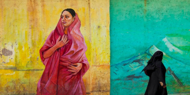 Local muslim woman dressed in a in black burqa is walking pass a wall painting in the city of Bangalore, India. The wall painting is in primary colors and shows a local woman in a red sari dress.