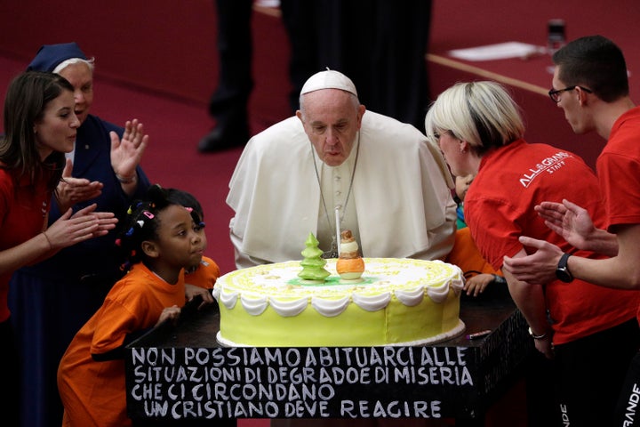 Pope Francis blows a candle atop of a cake he was offered on the eve of his 82nd birthday at the Vatican, Sunday, Dec. 16, 2018. The writing on the bottom of the cake reads: "We cannot get used to the situation of decay and poverty that surrounds us. A Christian must react."