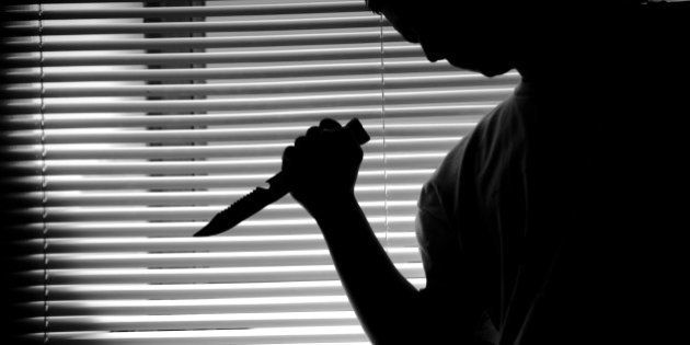 A silhouette of a teenager with a knife in his hand, in front of blinds in a dark room.