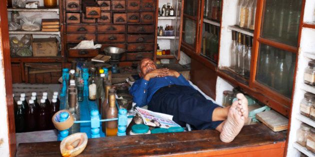 An Indian pharmacist sleeps in his small shop at midday. He is surrounded by cabinets and shelves stocked with drugs and ingredients he can make into medicines and lotions and potions using the pestle and mortar and scales