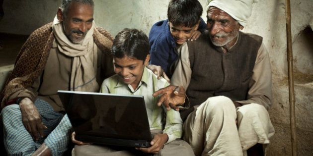 Happy village boy sitting cross-legged and using a laptop in the company of his father, grandfather and younger brother. This rural scene in India is taken beside an adobe wall with the grandfather's bamboo walking stick leaning against it in the background. The grandfather is pointing at the laptop and questioning his grandson, the father is looking with fascination at what his son is doing with the laptop and the boy's younger brother is peeking out from behind to see what is happening. This group portrait is shot at night with studio lights.