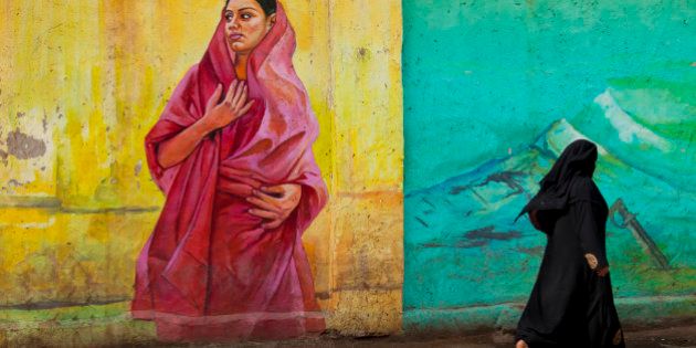 Local muslim woman dressed in a in black burqa is walking pass a wall painting in the city of Bangalore, India. The wall painting is in primary colors and shows a local woman in a red sari dress.