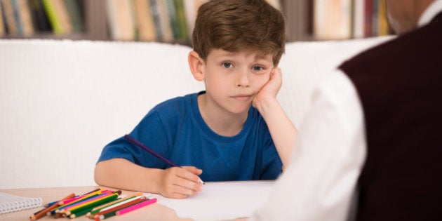 Sad little boy trying to draw in psychologist office. Focus on boy. There are many books in psychologist office