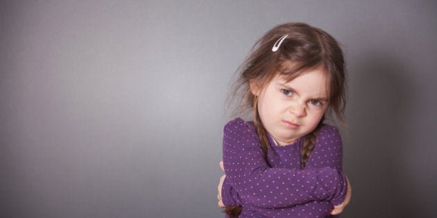 A little cute Girl is sulking. She is wearing a purple shirt in front of a gray background.