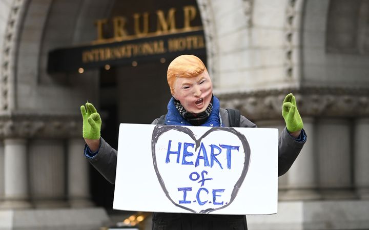 A man in a Donald Trump face mask poses in front of the Trump International Hotel during the march in Washington, D.C., wearing a sign that reads, "Heart of I.C.E."