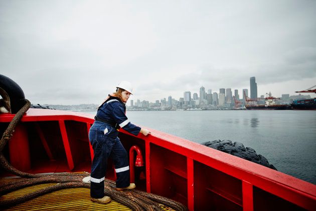 Female naval architect in hardhat and coveralls taking measurement along bulwark of commercial tugboat