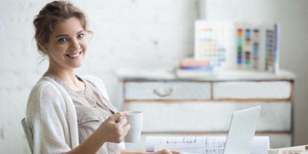 Portrait of beautiful happy smiling young designer woman sitting at home office desk with cup of coffee, posing, looking at camera. Attractive cheerful model using computer. Indoors