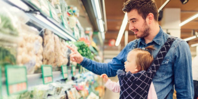 Father and his baby girl groceries shopping in local supermarket. He is carrying his daughter in baby carrier and choosing vegetable.