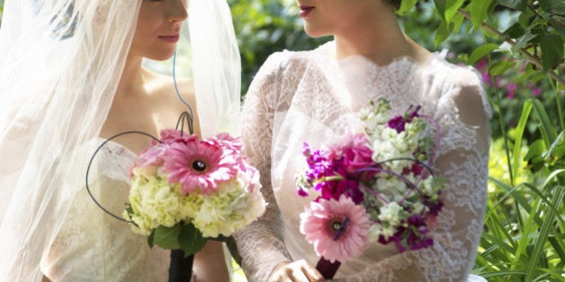 Horizontal outdoor garden shot of two young women in wedding dresses with bridal bouquets gazing into each others eyes.