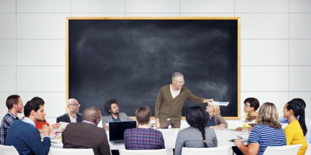 A gray haired man stands in front of a large blackboard and addresses a multiethnic group of students. The students are sitting around a large table.