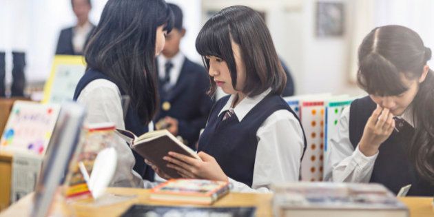 High school girl reading book in classroom. Female student is with classmates in brightly lit school. They are in uniforms.