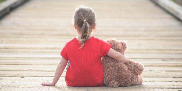 Little girl in a red dress waiting on a boardwalk hugging teddybear, view from behind