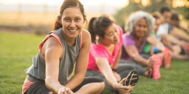 A group of adults attending a fitness class outdoors are doing leg stretches. The participants are arranged in a line. The focus is on a beautiful ethnic woman who is smiling toward the camera.