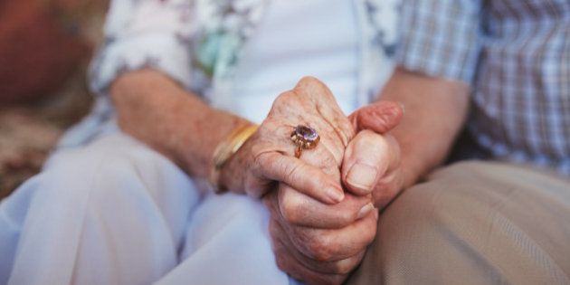 Cropped shot of elderly couple holding hands while sitting together at home. Focus on hands.