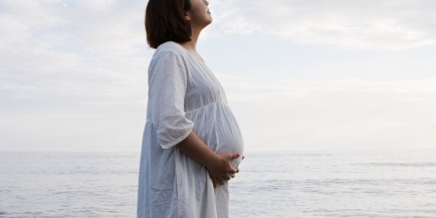 an asian expectant mother standing on the beach and looking up at sky with her eyes closed.