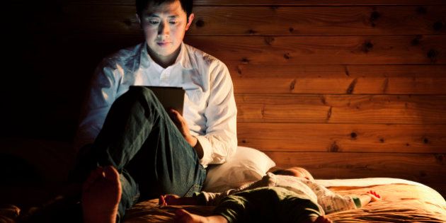 An asian father using a tablet pc on the bed at night, his baby boy sleeping by his side.