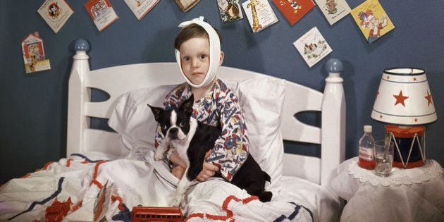 A sick boy with a bandage wrapped around his head sits in bed, a French Bulldog in his lap and surrounded by toys and get-well cards, 1950. There is a bottle of Benedryl and a lamp shaped like a toy drum on the nightstand next to him. (Photo by Lambert/Getty Images)