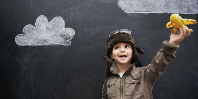 A little boy playing with a yellow toy airplane in front of clouds drawn on a blackboard