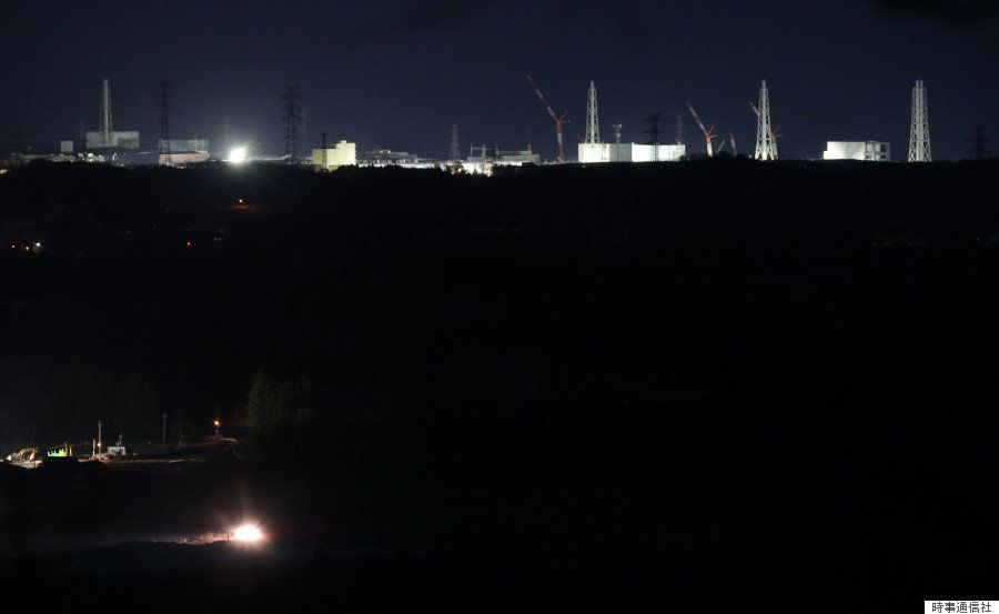 住民の避難が続き闇に包まれる町（下）と対象的に、ライトに照らされ浮かび上がる東京電力福島第一原子力発電所の建屋（1秒露光）。左下の明かりは車のライト（2016年03月10日夜、福島県富岡町から撮影）