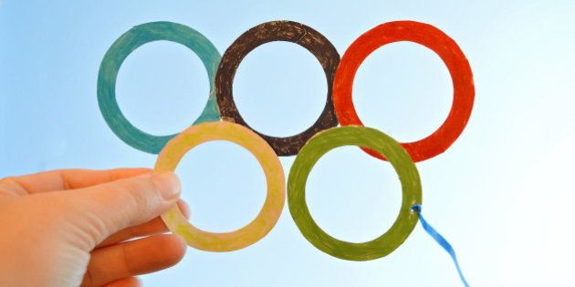 Child is holding a symbol for an Olympics, five Olympic circles made of colorful papers, cut out and colored, presenting the glasses.In the background is simple blue summer sky without any clouds.Sunny. warm day in the summer