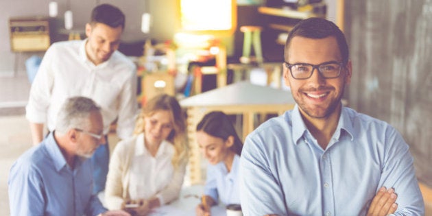 Handsome businessman in eyeglasses is looking at camera and smiling, in the background his colleagues are discussing work