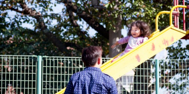 A little girl is sliding a slider at a park. She is happyly smiling. Her father is standing nearby and looking at her.