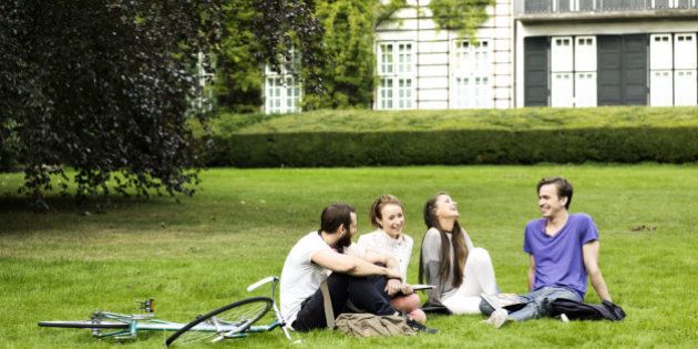 Four students sitting on the grass in a park, with some buildings in the background. Two female and two male students