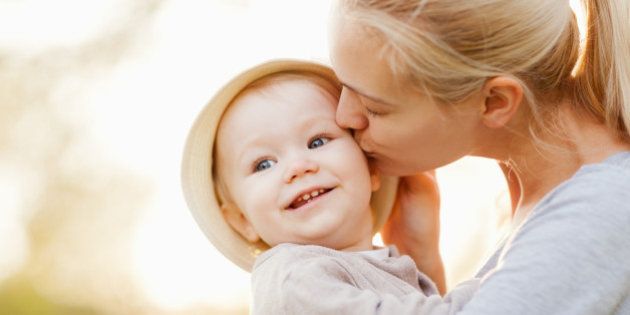Close up of a beautiful young mother with blonde hair kissing her one year old daughter on the cheek. The little girl is wearing a hat and a beige cardigan. Int he blurred background are some leafs on trees and a nice bright light.