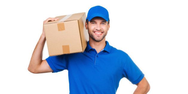 Joyful young courier carrying cardboard box on shoulder and smiling while standing against white background