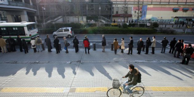 A man cycles past morning commuters waiting in line for a bus outside an East Japan Railways Co. train station after Tokyo Electric Co. announced the power outages in Urayasu city, Japan, on Monday, March 14, 2011. East Japan Railways Co., the nation's biggest rail company, was running only six of its busiest commuter lines and had stopped 32 other lines, according to its website. The railway's operational lines were running at 20 percent of their normal schedule. Photographer: Tomohiro Ohsumi/Bloomberg via Getty Images