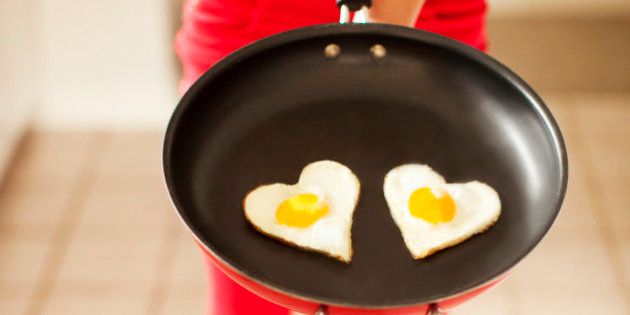 Woman dressed a red-patterend clothing holds a frying pan in front of her containing two heart-shaped eggs.