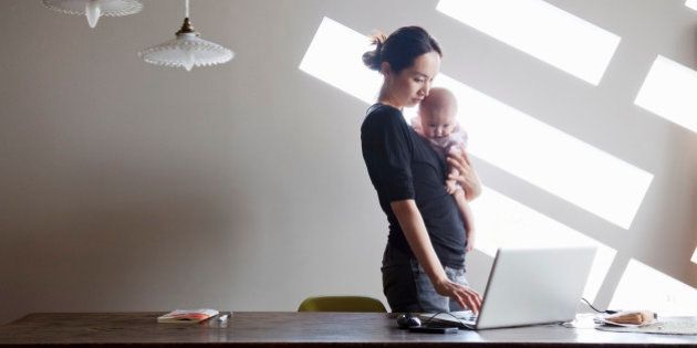 An asian gorgeous mother is standing and carrying her baby. She is working on laptop at home.