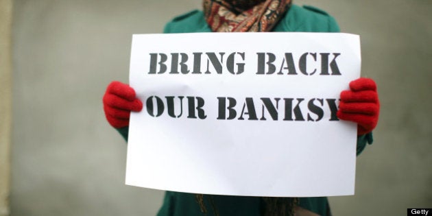 A woman holds a placard that reads 'Bring Back Our Banksy' during a protest next to a section of a wall where celebrated street artist Banksy's 'Slave Labour' graffiti artwork was removed in north London on February 23, 2013. The work that showed a young boy using a sewing machine to make the British flag has been carefully removed and will be auctioned in Miami where it's expected to fetch around 328,000 GBP (500,000 USD). Residents of the North London area have reacted angrily to the removal of the work, but the auction house says the piece was acquired legally. AFP PHOTO / JUSTIN TALLIS (Photo credit should read JUSTIN TALLIS/AFP/Getty Images)